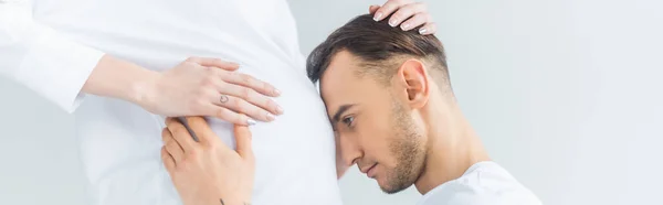 Вид сбоку молодого человека касающегося живота беременной жены изолированный на сером, панорамный снимок — стоковое фото