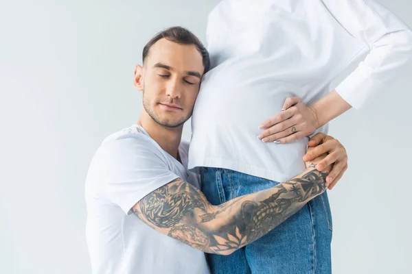 Seitenansicht des jungen tätowierten Mannes umarmt Bauch der schwangeren Frau mit geschlossenen Augen isoliert auf grau — Stockfoto