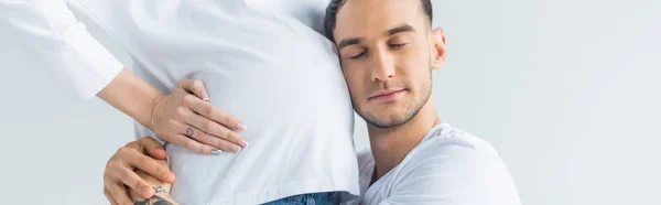 Side view of young tattooed man embracing belly of pregnant wife with closed eyes isolated on grey, panoramic shot — Stock Photo