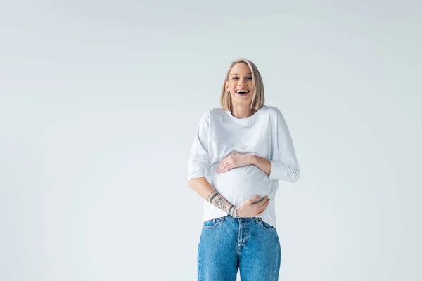 Happy young tattooed pregnant woman touching belly isolated on grey — Stock Photo