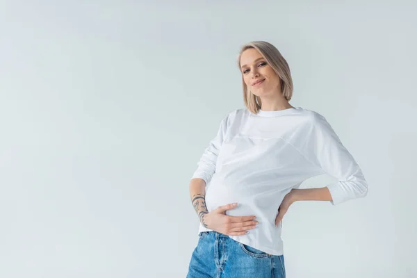 Lächeln junge tätowierte schwangere Frau berührt Bauch isoliert auf grau — Stockfoto