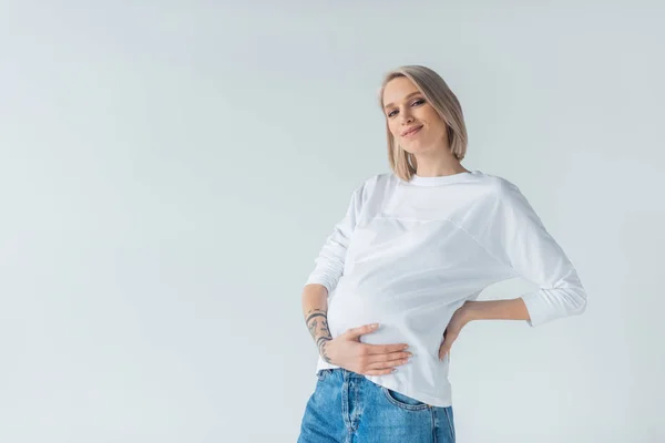 Smiling young tattooed pregnant woman touching belly isolated on grey — Stock Photo