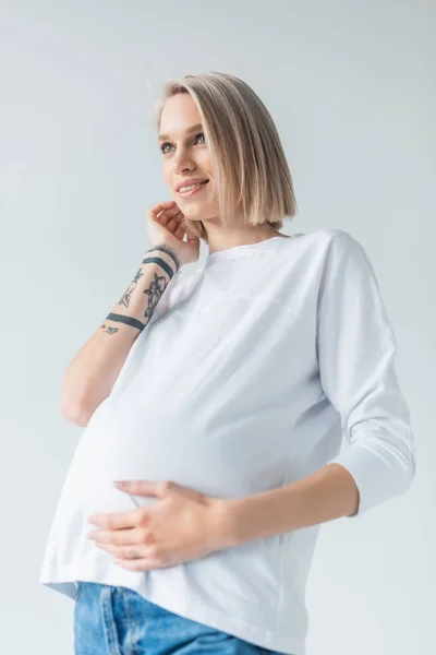 Smiling young tattooed pregnant woman touching belly isolated on grey — Stock Photo