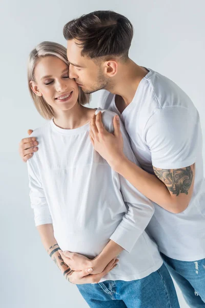 Young tattooed pregnant woman kissing with husband isolated on grey — Stock Photo