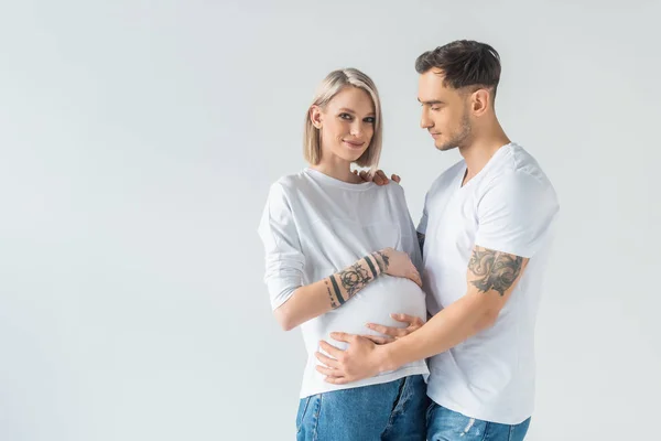 Sonriente joven mujer embarazada tatuada abrazándose con novio aislado en gris - foto de stock