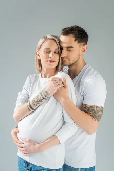 Joven mujer embarazada tatuada cerca de novio con los ojos cerrados aislados en gris - foto de stock