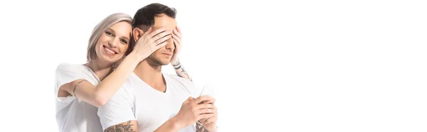 Souriant jeune fille tatouée couvrant les yeux de petit ami tandis que l'homme utilisant smartphone isolé sur blanc, panoramique shot — Photo de stock