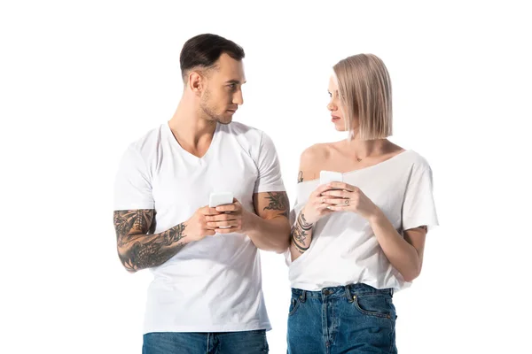 Young tattooed couple using smartphones and looking at each other isolated on white — Stock Photo