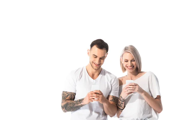 Sonriente joven pareja tatuada utilizando teléfonos inteligentes aislados en blanco - foto de stock