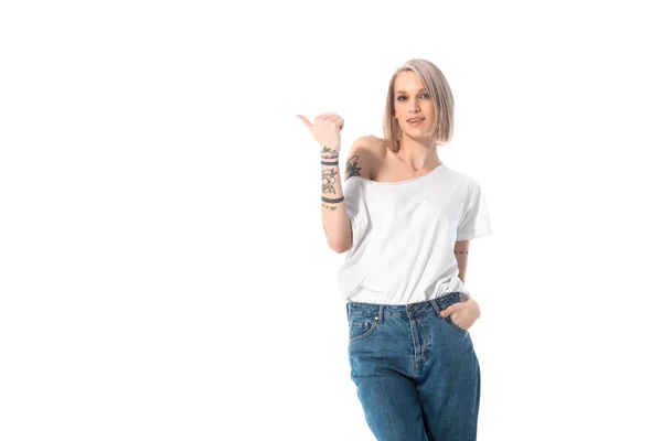Молода татуйована дівчина з рукою в кишені, вказуючи пальцем геть ізольована на білому — стокове фото