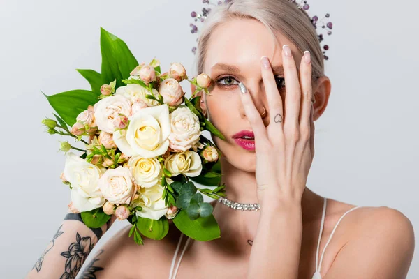Retrato de bela noiva tatuada posando com buquê floral e mão no rosto isolado em cinza — Fotografia de Stock