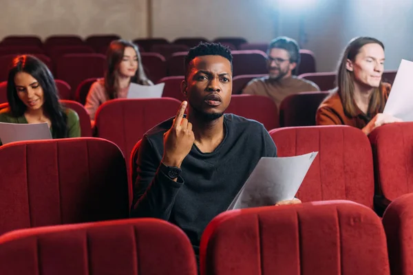Actor afroamericano ensayando y señalando hacia arriba mientras está sentado en sillas rojas con actores - foto de stock