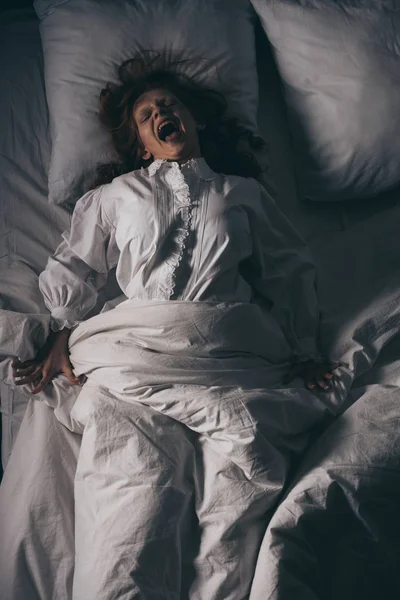 Вид зверху на демонічну злу дівчину в сорочці кричить, лежачи в ліжку — стокове фото