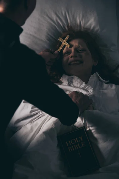 Exorcista con la biblia y cruz de pie sobre la chica demoníaca en la cama — Stock Photo