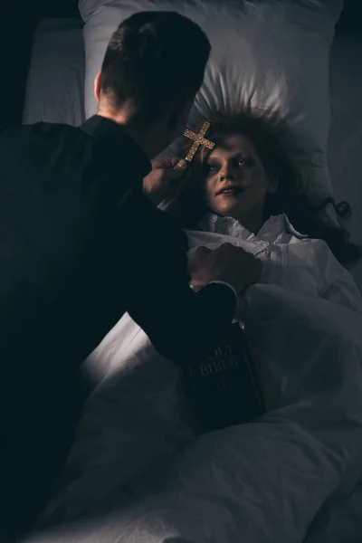 Exorcist with bible and cross standing over obsessed girl in bed — Stock Photo