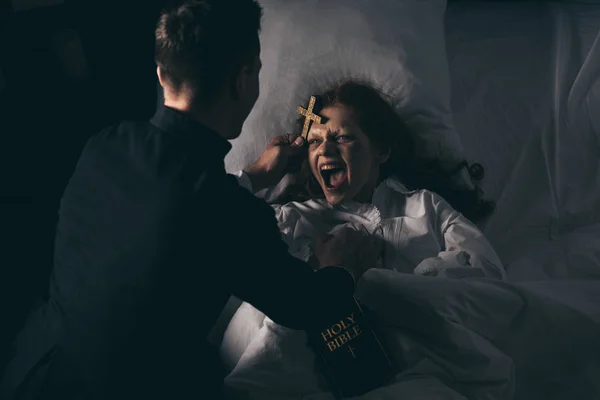 Macho exorcista con biblia y cruz de pie sobre demoniaca gritando chica en la cama - foto de stock