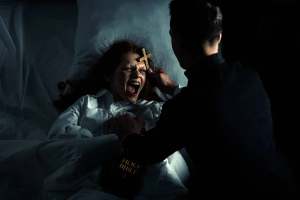 Exorcista con la biblia y cruz de pie sobre demoniaca gritando chica en la cama — Stock Photo