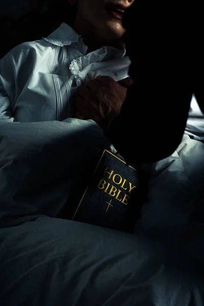 Обрізаний вид екзорциста з Біблією тримає руки з одержимою дівчиною в ліжку — стокове фото