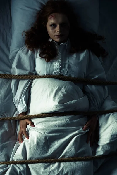 Одержимая женщина в ночной рубашке, связанная веревкой в постели — стоковое фото