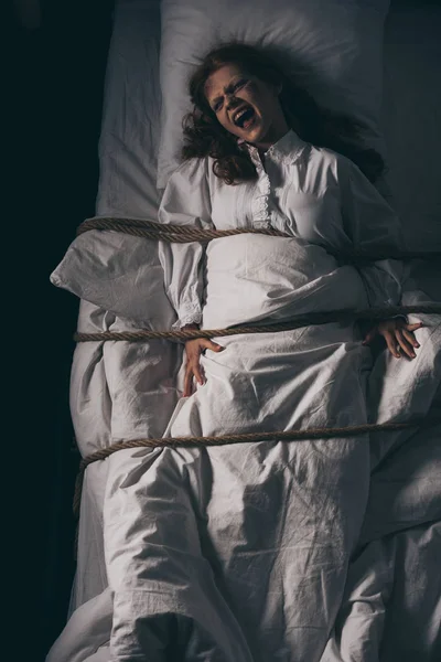 Creepy yelling girl in nightgown bound with rope in bed — Stock Photo