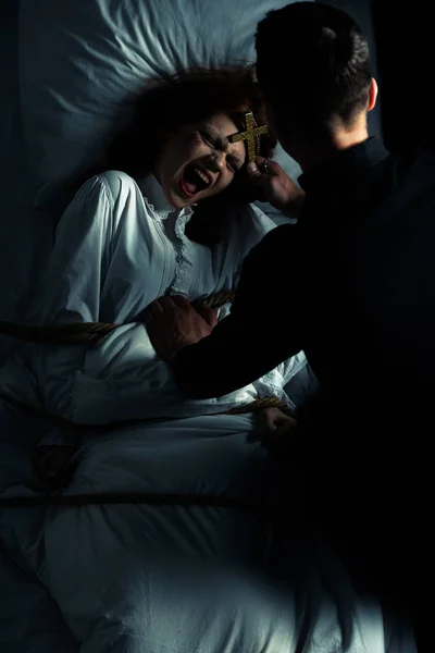 Exorcista sosteniendo cruz sobre demonio hembra atado con cuerda en la cama - foto de stock
