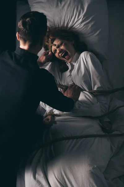 Exorcist holding cross over demonic obsessed girl bound with rope on bed — Stock Photo