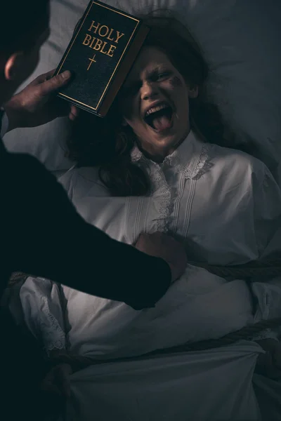 Exorcist with bible and obsessed screaming woman in bedroom — Stock Photo