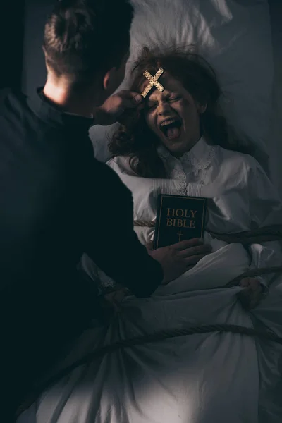 Exorcist with bible and cross standing over obsessed yelling girl in bed — Stock Photo