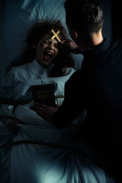 Exorcista masculino com bíblia e cruz em pé sobre demoníaco gritando menina na cama — Fotografia de Stock