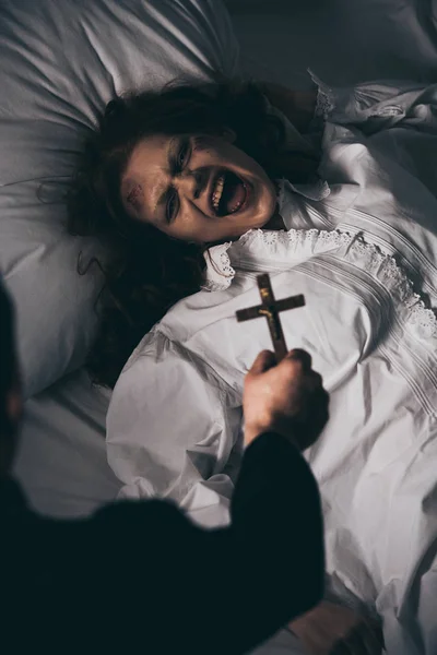 Exorcista sosteniendo cruz sobre espeluznante demonio gritando en la cama - foto de stock