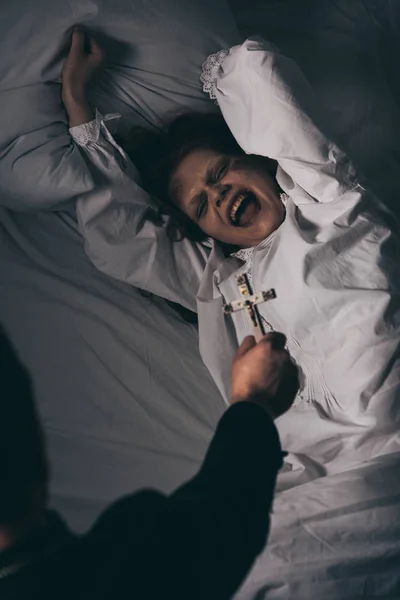 Exorcist holding cross over creepy screaming demon in bed — Stock Photo