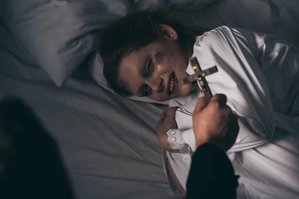 Exorciste tenant croix sur démoniaque obsédé souriant fille dans le lit — Photo de stock