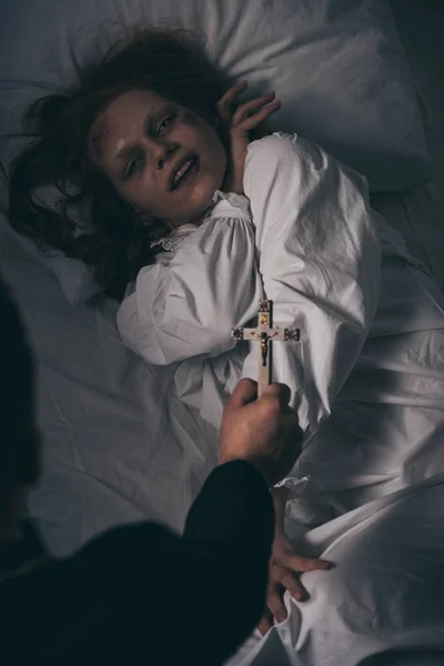 Exorcista sosteniendo cruz sobre demoníaca chica en cama - foto de stock