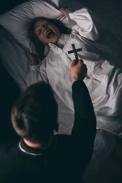 Exorcista sosteniendo cruz sobre obsesionado gritando chica en la cama - foto de stock