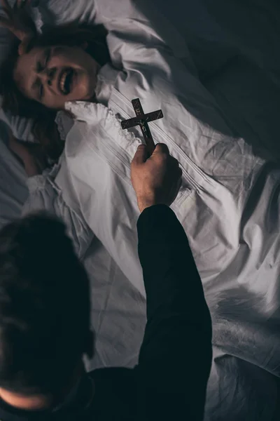 Exorcista sosteniendo cruz sobre obsesionado gritando chica en la cama - foto de stock