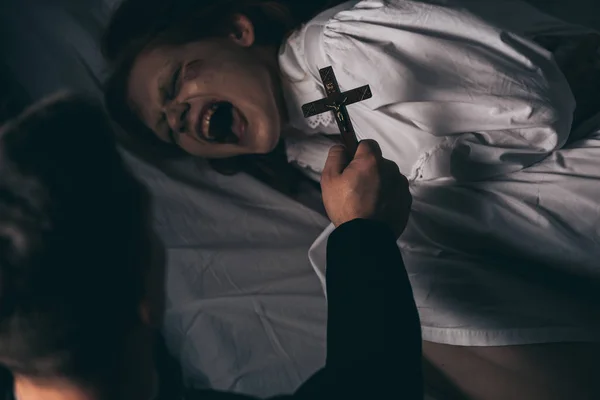 Exorcista sosteniendo cruz sobre demoníaca gritando chica en la cama - foto de stock