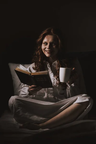 Demonic smiling woman in nightgown holding bible and cup on bed — Stock Photo