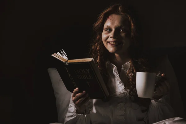 Демоническая улыбающаяся девушка в ночной рубашке, держащая библию и чашку на кровати — стоковое фото