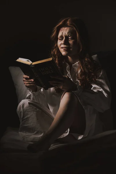 Демоническая плачущая женщина в ночной рубашке читает священную Библию на кровати — стоковое фото