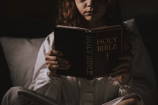 Cropped view of creepy girl in nightgown reading bible on bed — Stock Photo