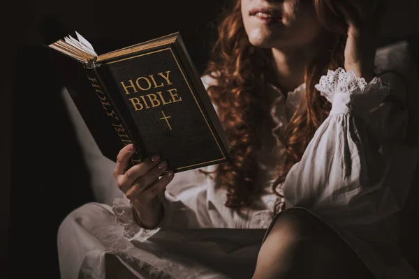 Обрезанный вид женщины-демона в ночной рубашке, читающей священную Библию на кровати — стоковое фото