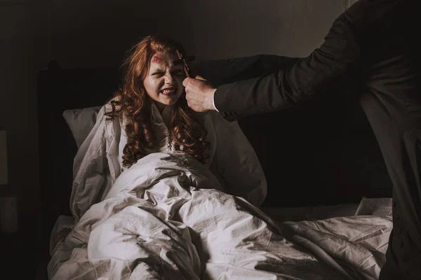 Exorcista sosteniendo cruz delante de chica obsesionada en la cama - foto de stock