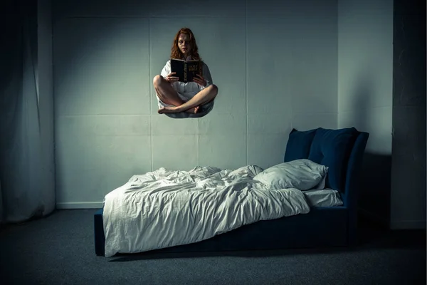 Демонічна жінка в нічній сукні вирівнюється над ліжком, читаючи Біблію — стокове фото