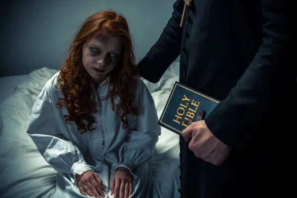 Exorcista sosteniendo la biblia y abrazando a chica demoníaca en el dormitorio - foto de stock