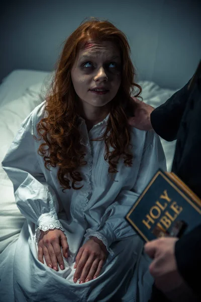 Екзорцист тримає Біблію і обіймає демонічну моторошну дівчину в спальні — стокове фото