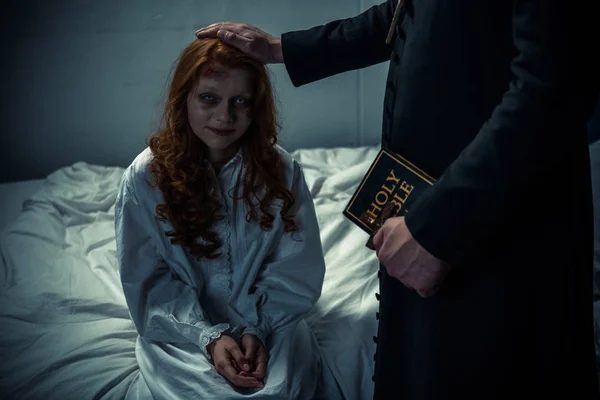 Exorcista sosteniendo la biblia y abrazando demoníaca espeluznante chica en el dormitorio - foto de stock