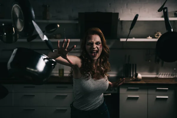 Demoniacal shouting girl gesturing with levitating kitchenware in kitchen — Stock Photo