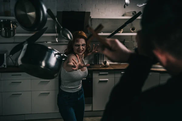 Selective focus of creepy demon with levitating cookware and exorcist with cross in kitchen — Stock Photo