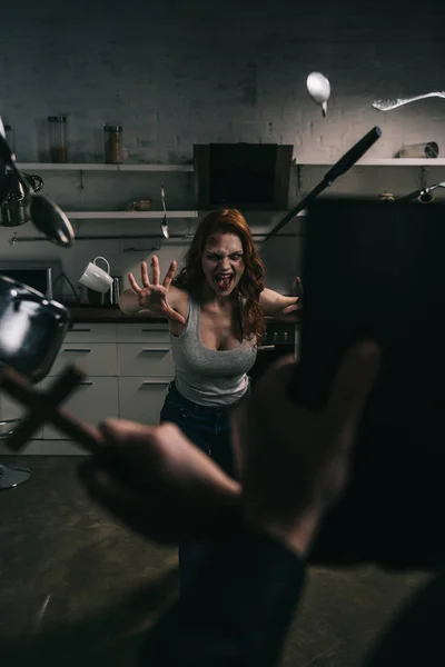 Selective focus of yelling demon with levitating cookware and exorcist with cross and bible in kitchen — Stock Photo