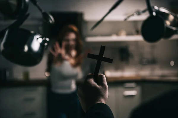 Selective focus of female demon with levitating cookware and exorcist with cross in kitchen — Stock Photo